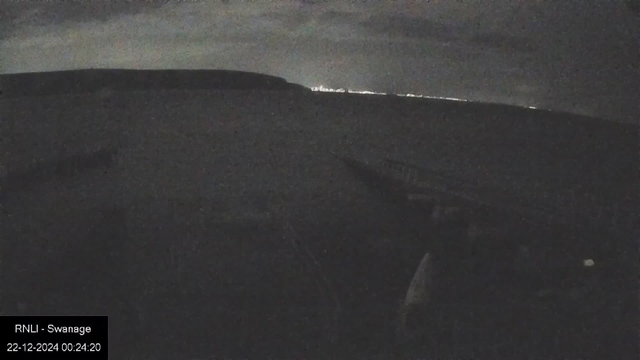 A nighttime view from a webcam showing a dark landscape. There is a silhouette of a hill or coastline in the distance beneath a cloudy sky. A row of faint lights lines the horizon, and in the foreground, a barely visible structure or pier extends into the scene. The bottom left corner has a label saying "RNLI - Swanage" with a timestamp of "22-12-2024 00:24:20".