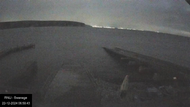 A dimly lit view from a webcam overlooking a body of water. In the foreground, a dock or pier extends into the water. The horizon is faintly visible with a line of distant lights reflecting on the water. The sky is overcast.