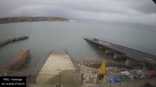 A seaside view at Swanage with a cloudy sky. The image shows a concrete boat ramp leading into calm water. Two piers extend into the sea on either side. In the foreground, there is a yellow kayak and several small boats on the rocky shore. Cliffs are visible in the distance across the water.