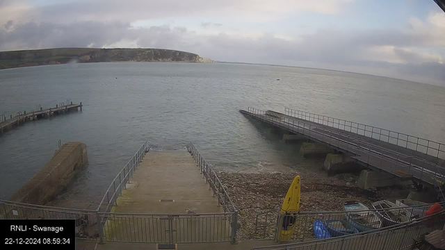 A view from a webcam shows a coastal scene with a boat ramp leading into the sea. There are two piers extending into the water on either side of the ramp. A rocky beach with some small boats and a yellow kayak are visible to the right. Distant cliffs line the horizon under a cloudy sky.