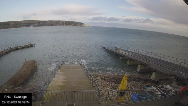 A scenic view from a webcam shows a coastal landscape with a calm sea stretching out to the horizon under a partly cloudy sky. In the foreground, a wide boat ramp leads to the water, flanked by metal railings. To the right, there is a concrete pier jutting into the sea. On the left, another shorter pier extends over the water. In the lower right corner, yellow surfboards or rescue boards are stored upright next to some small boats. In the background, a shoreline with grassy cliffs is visible. A time and date stamp in the lower left reads "22-12-2024 09:59:34," along with the text "RNLI - Swanage."