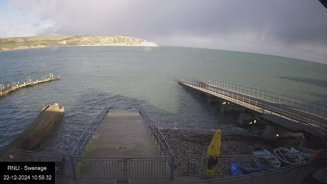 A coastal view from a webcam shows a seaside scene with a concrete slipway leading into the water, flanked by two piers. The ocean is calm with gentle waves, and the sky is overcast with patches of sunlight breaking through. In the foreground, there's a yellow kayak or surfboard, and several small boats are moored nearby. The distant shoreline features cliffs and green hills.