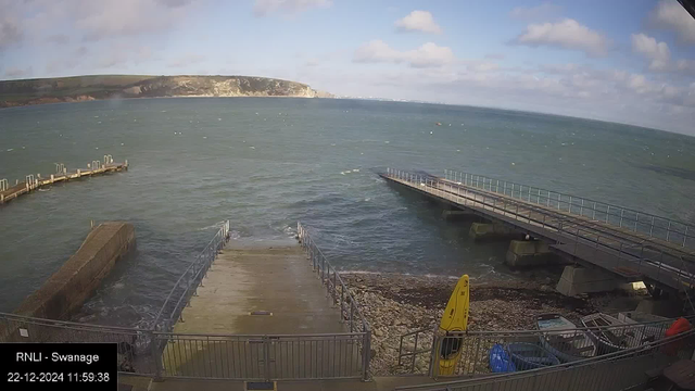 Webcam view of a coast in Swanage, showing a calm sea under a partly cloudy sky. In the foreground, there are two piers extending into the water and a concrete boat ramp leading to the sea. The coastline features cliffs and green hills in the distance. A yellow kayak is placed near the shore on a pebble-strewn beach. The timestamp on the image reads "22-12-2024 11:59:38."