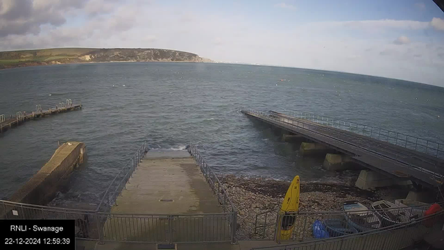 Webcam image showing a coastal view at Swanage. In the foreground, there is a concrete slipway leading into the sea, flanked by metal railings. To the right, there is a yellow kayak resting on a rocky shore beside some small boats. In the background, the sea extends to the horizon with a small pier to the left and a bridge to the right. Cliffs and grassy hills are visible in the distance under a cloudy sky. The time and date stamp reads "22-12-2024 12:59:39."