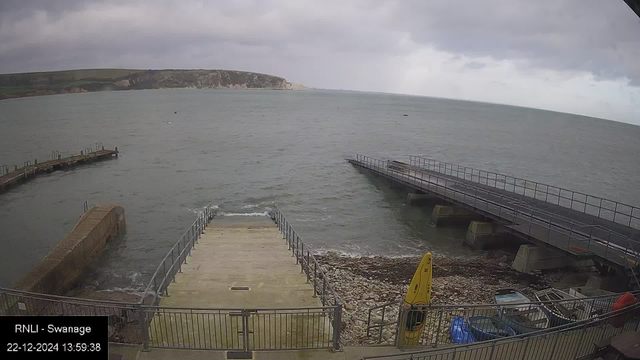 A web camera image of a coastal scene under a cloudy sky. The foreground features a boat ramp extending into the sea, flanked by safety railings. On the left, a concrete pier projects into the water. On the right, another dock stretches over the water, supported by pillars. A rocky shoreline is visible, with a few small boats and a yellow kayak on the right side. In the background, grassy cliffs form a horizon above the sea.